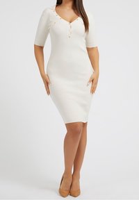 Vestito bianco a coste con maniche corte, scollatura a V profonda e bottoni decorativi. Silhouette aderente che arriva fino al ginocchio, abbinato a tacchi color beige.