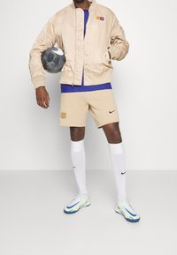 Beige Bomberjacke, blaues Hemd, beige Shorts mit Logo, weiße Socken und blau-weiße Fußballschuhe. Hält einen schwarzen Fußball.
