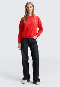 Rode sweatshirt met een ronde hals en geborduurd logo, gecombineerd met donkerblauwe jeans en witte sneakers. Casual pasvorm en gladde textuur.