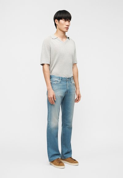 Άνδρας όρθιος, φοράει πλεκτό πόλο με κοντά μανίκια ανοιχτού γκρι χρώματος, ξεθωριασμένο μπλε τζιν και καφέ σουέτ αθλητικά παπούτσια σε απλό λευκό φόντο.