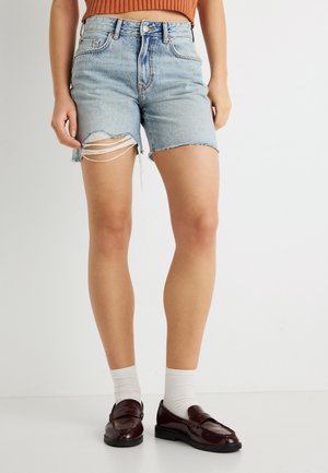 Hellblaue Jeansshorts mit ausgefranstem Saum und seitlichen Ausschnitten, mit hoher Taille und Standard-Fünf-Taschen-Design, kombiniert mit burgunderfarbenen Loafers.