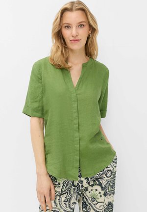 Lyshåret kvinde iført en grøn kortærmet bluse og mønstret creme- og marineblå blomstrede bukser, stående mod en hvid baggrund.