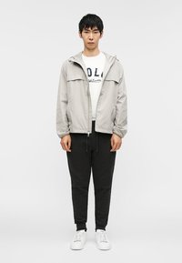 Ανοιχτό γκρι ελαφρύ μπουφάν με κουκούλα, εμπρός φερμουάρ και ελαστικές μανσέτες, φορεμένο πάνω από λευκό μπλουζάκι με σχέδιο και μαύρη φόρμα.