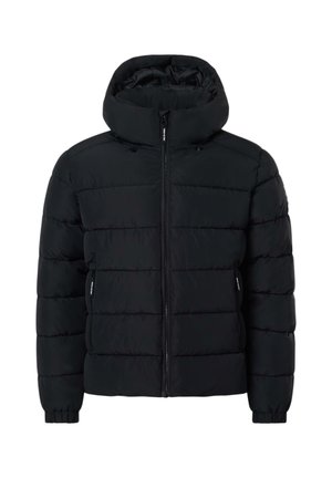 Jeune homme portant une veste noire matelassée à capuche et un jean bleu, regardant par-dessus son épaule sur un fond clair uni.
