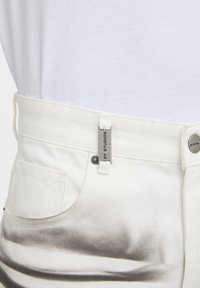 Jean en denim blanc avec un effet de tache grise dégradée près de la poche avant et une étiquette de marque en métal sur la passants de ceinture.