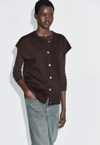 Kobieta ubrana w ciemnobrązową kamizelkę zapinaną na guziki, na długim rękawie, w jasnoniebieskich dżinsach, stojąca z jedną ręką w kieszeni, oparta o białą ścianę.