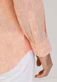 Camisa a rayas en naranja y blanco, con puños de botón, tela ligera y una textura sutil. El dobladillo es más corto a los lados.