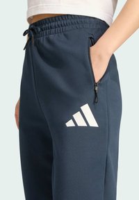 Námornícky modré športové nohavice s viazaním v páse, bočnou zipsovou kapsou a výrazným bielym trojprúžkovým logom na stehne.