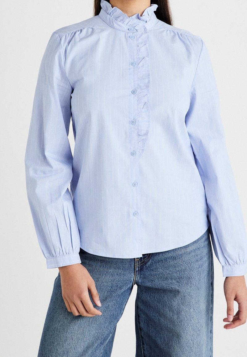 Femme portant un chemisier bleu clair à manches longues avec une patte de boutonnage à volants et un col, associé à un jean en denim bleu.