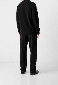 Pull en maille côtelée noire à manches longues, associé à un pantalon noir ajusté et des chaussures noires, vu de dos.