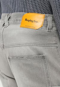 Ανοιχτό γκρι τζιν denim με ένα φωτεινό πορτοκαλί δερμάτινο εμβλημα Replay Inc. στη μέση πίσω, φορεμένα με γκρι πλεκτό πουλόβερ.