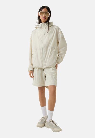 Light beige windbreaker with a zip and two pockets, paired with matching shorts. Chunky beige sneakers and oversized light sunglasses.
