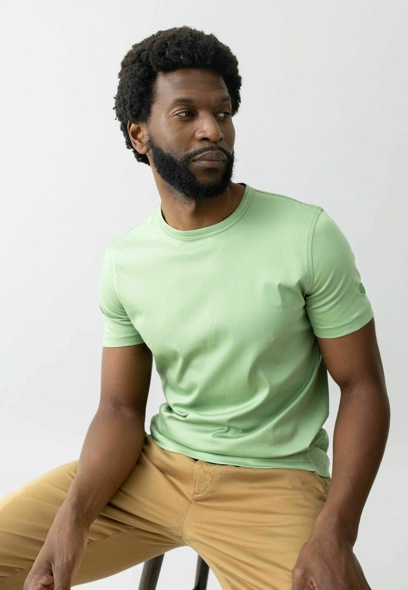 Menta zöld, rövid ujjú póló, kerek nyakkivágással és diszkrét ujjtartással, világos bézs nadrággal párosítva. Sima szövet textúra.