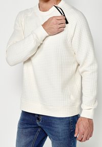 Felpa leggera di colore crema con motivo trapuntato, colletto e polsini a costine, caratterizzata da una finitura testurizzata. Abbinata a jeans in denim blu.