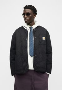 Άνδρας που φορά γυαλιά ηλίου, μαύρο μπουφάν με καπιτονέ επένδυση πάνω από λευκή φούτερ με κουκούλα, μπλε γραβάτα με ρίγες και σκούρο παντελόνι, στέκεται μπροστά σε ένα απλό λευκό φόντο.