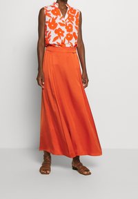 Ärmelloses Oberteil mit Blumenmuster in Weiß und Orange, mit Kragen, kombiniert mit einem langen, fließenden orangefarbenen Satinsrock und braunen Riemchensandalen.