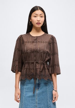 Vrouw met lang zwart haar die creolen draagt, een bruine blouse met oogjes en een strik in de taille, en een blauwe spijkerrok tegen een effen achtergrond.