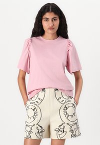 T-shirt rose clair à manches courtes avec des détails froncés sur les épaules, associé à un short crème avec un motif abstrait noir.