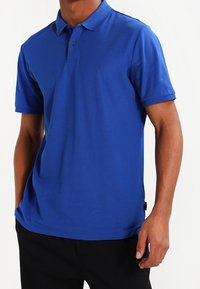 Man som bär en enfärgad kortärmad blå pikétröja med krage och två knappar, i kombination med svarta byxor.