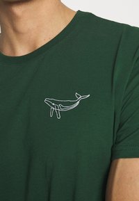 Camiseta de algodón verde con un diseño de ballena blanca bordado en el pecho, mangas cortas y escote redondo.