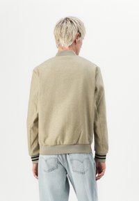 Beige bomber jacket made of woven fabric, featuring a ribbed collar and cuffs with black stripes. Back view shows a streamlined design.