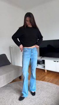 Vrouw die lichtblauwe jeans aanpast, draagt een zwarte top met lange mouwen en zwarte schoenen in een woonkamer met een bank en tv.