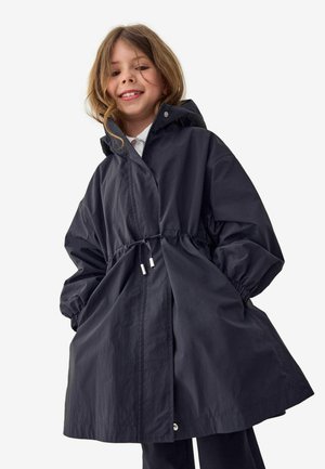Uśmiechnięte dziecko, ubrane w luźną ciemną kurtkę z kapturem, elastycznymi mankietami i sznurkiem w pasie, ręce w kieszeniach, na jednolitym tle.