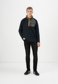 Pullover in pile blu navy con tasca sul petto verde, cerniera arancione e jeans skinny neri. Il modello indossa sneakers nere con strisce bianche.