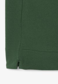 Gros plan d’un tissu tricoté vert montrant un ourlet avec une petite fente latérale et des détails visibles de couture.