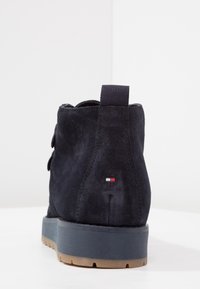 Marinblå mocka ankelskål med en texturerad yta, med en dragflik och en liten logotyp i röd, vit och blå på hälen. Gummisula.