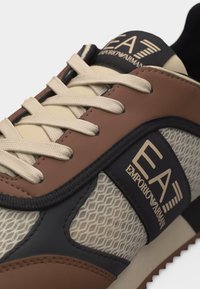 Κοντινό πλάνο καφέ, μπεζ και μαύρου αθλητικού παπουτσιού με μπεζ κορδόνια και το λογότυπο EA Emporio Armani σε δίχτυ και δερμάτινα πάνελ.