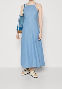 Vaaleansininen denimmaksimekko ohuilla olkaimilla, vartalonmyötäisellä yläosalla ja levenevällä hameella. Yhdistettynä kudottuun sinivalkoiseen laukkuun ja valkoisiin sandaaleihin.