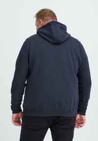 Námornícky modrá mikina vyrobená z mäkkej tkaniny, s kapucňou na šnúrku, dvoma bočnými vreckami a rebrovanými manžetami a lemom. Zobrazený pohľad zozadu.