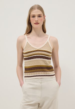 Jonge vrouw met lang blond haar, gekleed in een mouwloze gehaakte top met beige, bruine en gele horizontale strepen en lichtgekleurde broek.