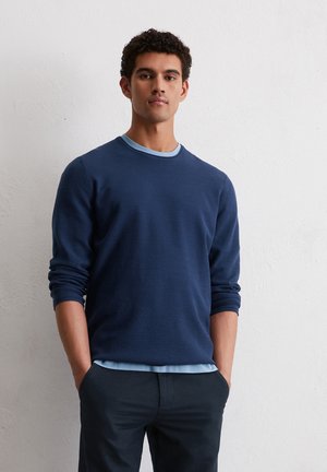 Dunkelblauer strukturierter Pullover mit einem hellblauen Rundhalsausschnitt. Das Model hat die Hände in den Taschen und trägt ein schichtenförmiges hellblaues Hemd darunter.