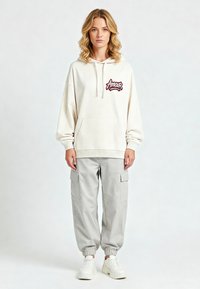 Felpa crema con grafica "Amour", tasca a marsupio e cappuccio con cordino. Abbinata a pantaloni cargo grigi chiari e sneaker bianche.