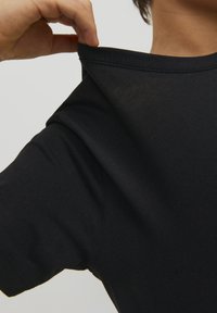 T-shirt di cotone nero con scollo rotondo, che mostra la texture liscia del tessuto e una leggera elasticità; il colletto è sollevato da una mano.