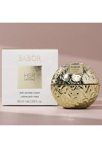 Kultainen pallomainen purkki, jossa on teksturoitu pinta, valkoisen laatikon vieressä. Laatikossa kohopainettuja kirjaimia. Molemmat kertovat ryppyjä ehkäisevästä voiteesta, 50 ml.