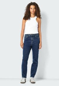 Modri kavbojki s ravnim krojem, visokim pasom in sprednjimi žepi, kombinirani z belo brezrokavno majico in sivimi supergami.