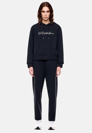 Giovane donna che indossa una felpa nera con cappuccio e scritta bianca, pantaloni neri e scarpe da ginnastica bianche, in piedi davanti a uno sfondo chiaro e uniforme.