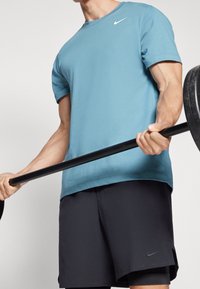 Osoba v modrém tričku Nike a černých šortkách Nike zvedá činku s černými závažími na jednobarevném pozadí.