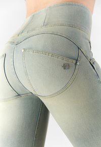 Jeans di un blu chiaro sbiadito con una vestibilità aderente, caratterizzati da una leggera lucentezza, due tasche posteriori e un piccolo logo metallico del brand sulla tasca destra.