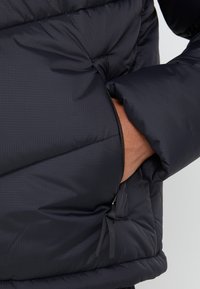 Giacca a vento nera con tasca con zip e tessuto testurizzato con un delicato motivo a griglia. Il design presenta una finitura liscia e isolante.
