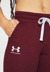 Burgundské tepláky s bílým šňůrkem, boční kapsou a bílým logem Under Armour na levém stehně mají měkkou texturu.
