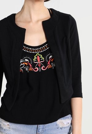 Kobieta nosząca czarny krótki sweterek na czarnej bluzce z kolorową kwiatową haftem i jasnoniebieskie, podarte dżinsy.