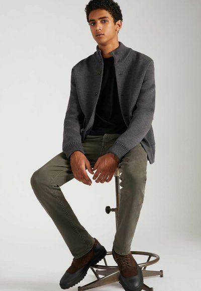 Veste en tricot grise avec un col haut, chemise noire, pantalon vert olive et chaussures à lacets marron. Modèle assis sur un tabouret de bar.