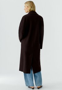 Lange donkerbruin wollen jas met een rugsplit. Heeft een opstaande kraag en een ontspannen silhouet, gecombineerd met lichtblauwe wijde spijkerbroeken.