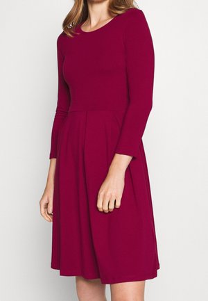 Kobieta w bordo sukience z długim rękawem, sięgającej do kolan, dopasowanej, z plisowaną spódnicą, stojąca na tle jednolitego jasnego tła.