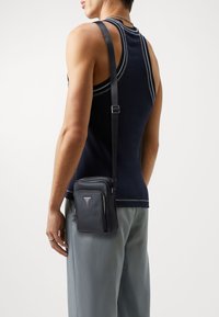 Bandolera de cuero negro con un acabado texturizado, bolsillos con cremallera y un logo triangular. Lleva puesta sobre un pantalón gris con una camiseta de tirantes oscura.