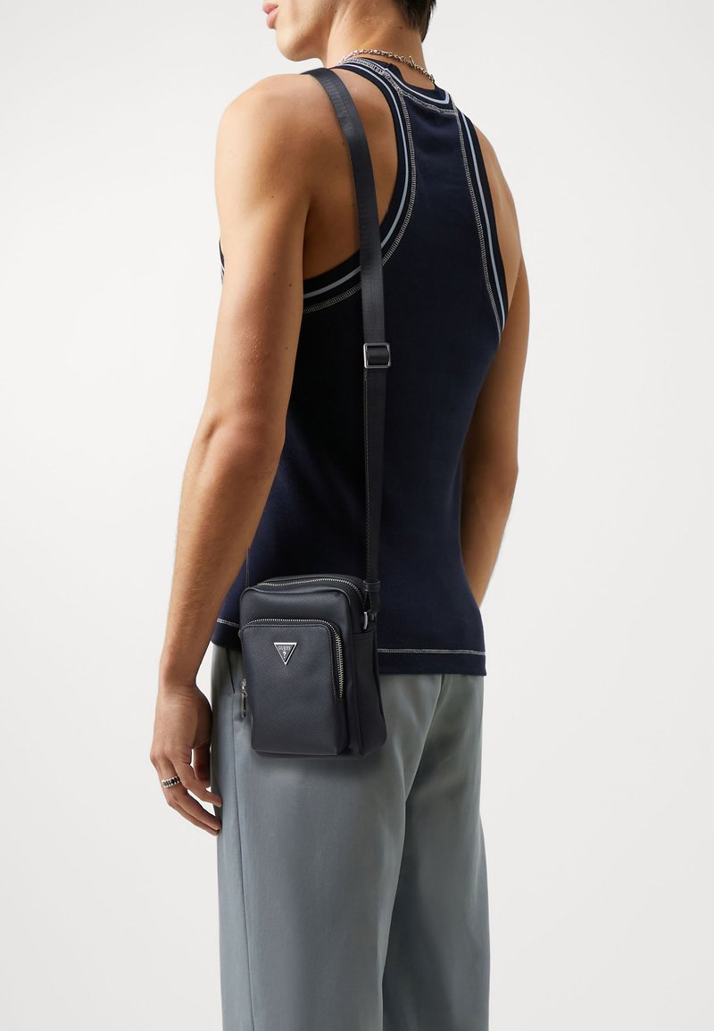 Bandolera de cuero negro con un acabado texturizado, bolsillos con cremallera y un logo triangular. Lleva puesta sobre un pantalón gris con una camiseta de tirantes oscura.